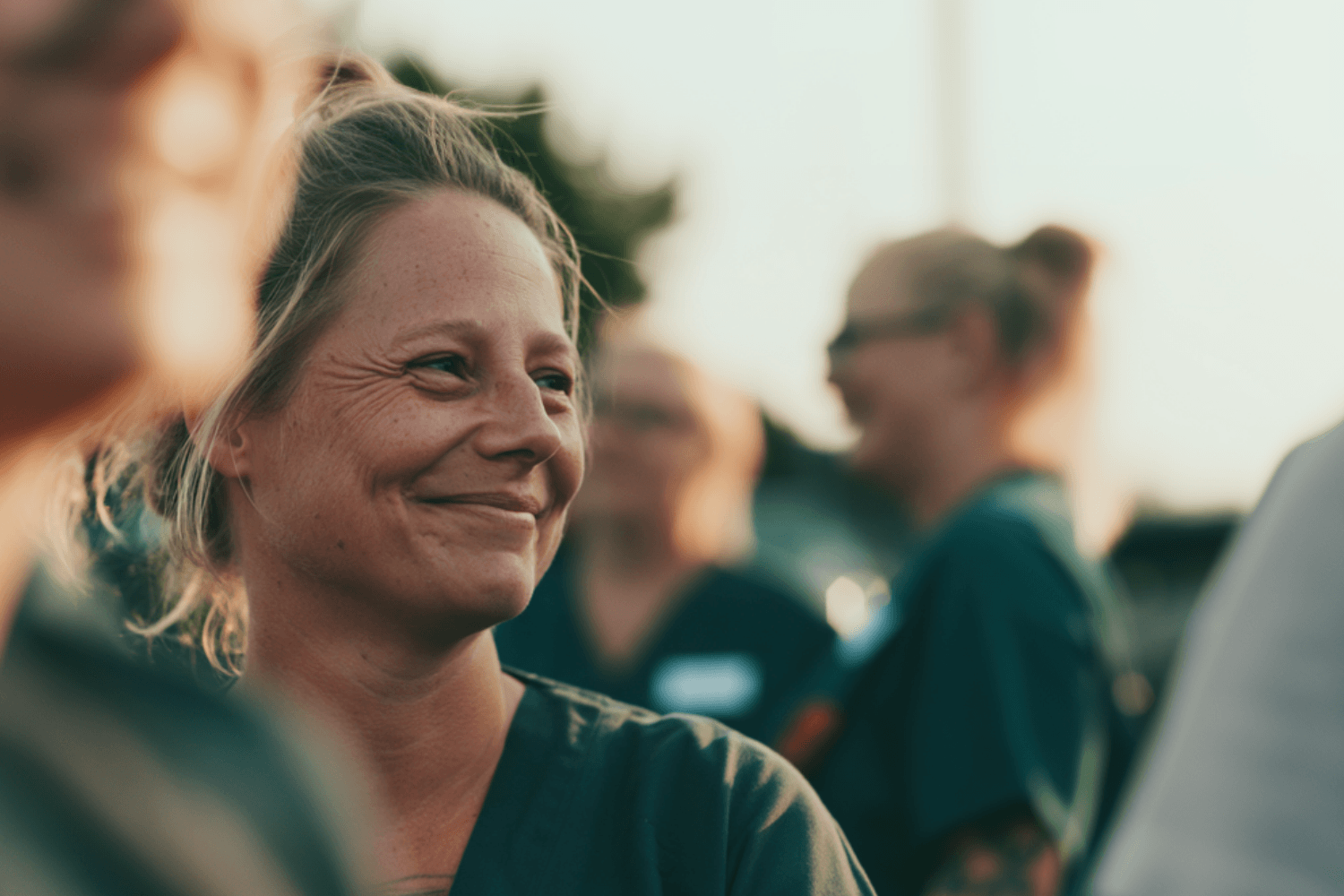 Eine blonde Gesundheits- und Krankenpflegerin steht mit Kolleg*innen im Freien während der Pause und sieht zufrieden und fröhlich aus